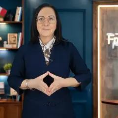 Kobieta w okularach i granatowej sukience, z rękami złożonymi w romb, na tle regału z książkami i neonu z napisem "Francuski Biegle".