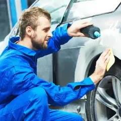 Mechanik w niebieskim kombinezonie nakłada szpachlę samochodową, czekając aż wyschnie, by naprawić karoserię.