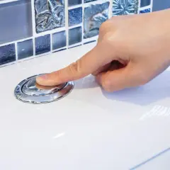Dłoń naciska chromowany przycisk spłuczki WC na białej ceramice, w tle mozaika.