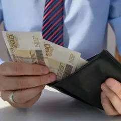 Mężczyzna w koszuli i krawacie wkłada banknoty 200 zł do portfela. Może to być wypłata, premia lub odprawa pośmiertna.
