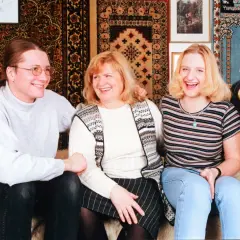 Trzy uśmiechnięte osoby siedzą na kanapie: mężczyzna z długimi włosami i kobieta w środku, a obok nich młoda kobieta.