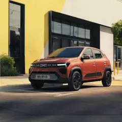 En orange Dacia står parkerad vid en laddstation framför en modern byggnad. Är Dacia bra eller dålig? Denna ser lovande ut!
