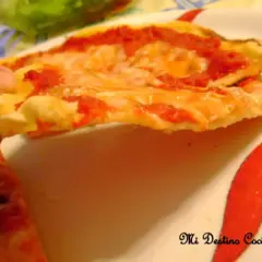 Una mano sostiene una rebanada de pizza con queso derretido y salsa de tomate, sobre un plato decorado con pimientos rojos. La masa, hecha con semola de trigo, luce dorada y crujiente.