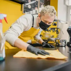 Barista con mascherina e guanti effettua la pulizia della cucina del ristorante, igienizzando il bancone con un panno giallo.