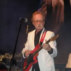 Muzyk w białej marynarce gra na czerwonej gitarze. Na scenie widać też perkusję i klawisze.
