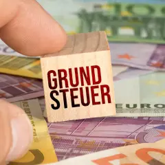 Ein Finger hält einen Holzstein mit der Aufschrift "GRUNDSTEUER" über Euro-Banknoten. Der **Grundsteuer Messbetrag** ist hier visuell dargestellt.