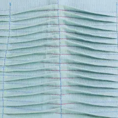 Close-up of light blue fabric with rows of pintucks, creating a textured, pleated effect. Red and blue threads highlight the stitching.