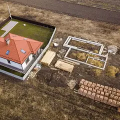 Zabudowa zagrodowa w budowie: gotowy dom z czerwonym dachem, fundamenty nowego budynku, drewno i palety cegieł.