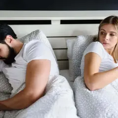 Ein Mann liegt im Bett und schaut auf sein Handy, während seine Frau ihn misstrauisch beobachtet. Er guckt vielleicht gerade Porno.