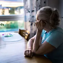 Starsza kobieta siedzi przy stole, zmartwiona. Zima i odcięcie prądu sprawiają, że czuje się samotna i bezradna.
