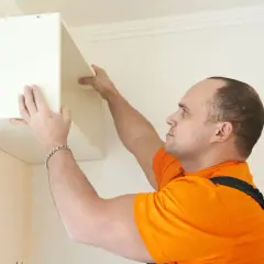 Un homme en orange est en train de fixer un meuble lourd au mur. Il positionne soigneusement l'étagère avant de la visser.