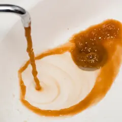 Eau brune et trouble coule d'un robinet dans un lavabo blanc, laissant des traces rouille. On dirait du poison dans l'eau.