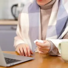Osoba owinięta kocem, z chusteczką w dłoni, pracuje przy laptopie. Obok kubek i zużyte chusteczki. Może to być sytuacja związana z art. 148 kp.