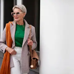 Elegancka kobieta po 60. w stylowej marynarce w kratkę, zielonej bluzce i białych spodniach. Pomarańczowy szal dodaje jej uroku.