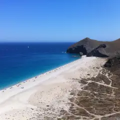 Playa de arena blanca y mar azul intenso, con acantilados rocosos y vegetación árida. Un velero navega en la distancia, un capricho del paisaje.
