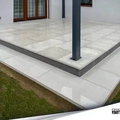 Nowoczesny taras z płyt betonowych, z podwyższonym stopniem i schodkami, otoczony zieloną trawą.