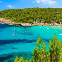 Bahía de aguas turquesas y cristalinas, con veleros y lanchas navegando. Al fondo, una playa de arena blanca y una frondosa vegetación que llega hasta la cala pudent.