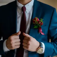 Elegancki mężczyzna w granatowym garniturze, białej koszuli i bordowym krawacie. Na klapie marynarki ozdobna butonierka z czerwonych róż.