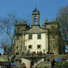 Edificio religioso con campanario y cruz. Gente en el balcón y en la plaza. Un árbol desnudo en primer plano.