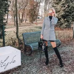 Elegancka kobieta w płaszczu w pepitkę, przypominającym kurzą stopkę, pozuje w parku.