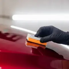 Ręka w czarnej rękawiczce nakłada preparat na błyszczący, czerwony lakier samochodu. Efekt jest olśniewający, niczym **kolor perłowy**.
