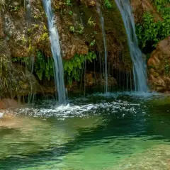 Cascada de la Osera, con agua cayendo sobre rocas cubiertas de musgo y vegetación exuberante, formando un estanque de aguas cristalinas.