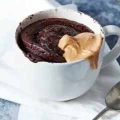 Schokoladiger high protein tassenkuchen mit Erdnussbutter-Topping in einer weißen Tasse, daneben ein Löffel.