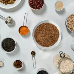 Różne rodzaje ziaren i nasion, w tym fasola, soczewica, ryż, gryka, ciecierzyca i rodzynki, ułożone na stole. Jedna łyżka stołowa ma ok. 15 ml.