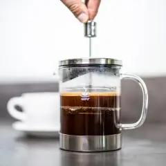 Eine Hand drückt den Stempel herunter, um Kaffee im Kaffeebereiter zuzubereiten. Daneben steht eine weiße Tasse.