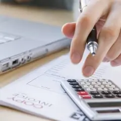 Dłoń z długopisem nad kalkulatorem i fakturą, obok laptop. Analiza pozostałych kosztów operacyjnych.