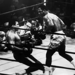 Joe Frazier fights in the ring, delivering a powerful blow as his opponent stumbles. The crowd watches intently.