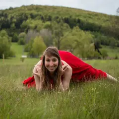 Kobieta w czerwonej sukni wykonuje ćwiczenia gimnastyki słowiańskiej na trawie, zalesione wzgórze w tle.