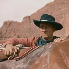 John Wayne w kapeluszu, oparty o konia, w krajobrazie przypominającym miasto bezprawia.