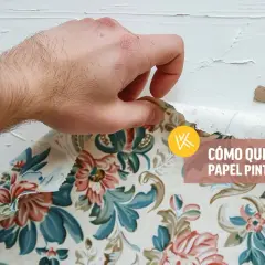 Mano retirando papel pintado floral de una pared blanca.
