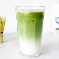 En snygg, isig matcha latte med lager av mjölk och grönt te. Bredvid står en visp för att göra matcha.