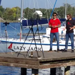 Dwóch mężczyzn na pomoście nagrywa film na tle żaglówki "Lazy Jack". Na pomoście leży kot.
