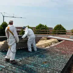 Pracownicy w kombinezonach usuwają eternit z dachu. W tle widać żuraw budowlany i drzewa.