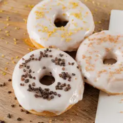 Drei Donuts mit weißer Glasur und Streuseln aus Schokolade, Zucker und Zitrone liegen auf einem Holzbrett.