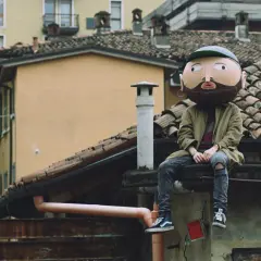 Un personaggio con una maschera gigante, simile a un cratere, siede sui tetti. I testi di Frah Quintale potrebbero raccontare la sua storia.