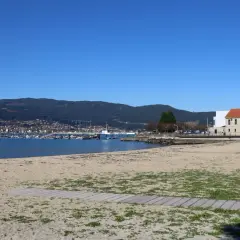 La playa de arealonga, con arena dorada y vegetación dispersa, se extiende hacia un mar azul tranquilo. Al fondo, un puerto deportivo con barcos y una ciudad en la ladera de una montaña verde.