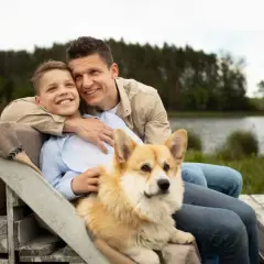 Ojciec i syn siedzą przy jeziorze z psem. Corgi kładzie się na kolanach chłopca, bo czuje się bezpiecznie i kochany.