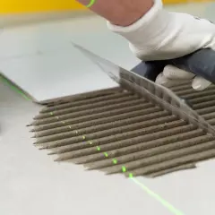 Ręka w rękawiczce rozprowadza zaprawę klejową kielnią po podłodze.