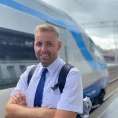 Uśmiechnięty mężczyzna w białej koszuli i niebieskim krawacie, gotowy do pracy. To on prowadzi pociąg, który stoi obok na torach.