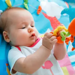 Niemowlę leżące na kolorowej macie bawi się pomarańczową zabawką.
