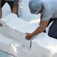 Dwóch budowlańców układa białe bloczki z perlitu, tworząc fundament. Jeden z nich w hełmie precyzyjnie dopasowuje element.