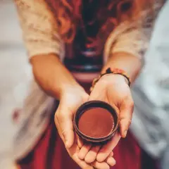 Dłonie trzymają małą, glinianą miseczkę z ciemnobrązowym płynem, przypominającym napój ceremonialny, być może ayahuasca lub DMT.