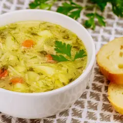 Aromaticzna, tradycyjna zupa jarzynowa z kawałkami mięsa, marchewką i ziemniakami, ozdobiona natką pietruszki. Obok kromki chleba.
