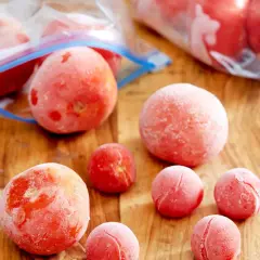 Gefrorene Tomaten liegen auf einem Holzbrett. Einige sind in Gefrierbeuteln, andere einzeln. Perfekt für Soßen und Suppen, wenn man Tomaten einfrieren möchte.