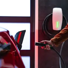Ręka trzymająca ładowarkę do samochodu elektrycznego przy czerwonym aucie. Obok widać biały wallbox z zielonym wskaźnikiem.
