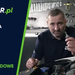 Mężczyzna pokazuje żarówkę samochodową, przygotowując się do jej wymiany. W tle warsztat z narzędziami.